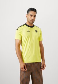 Kieta rankovė geltona sportinė marškinė su juodais akcentais, turinčiais apvalią apykaklę, Adidas logotipą ir juodą liūto grafiką priekyje.