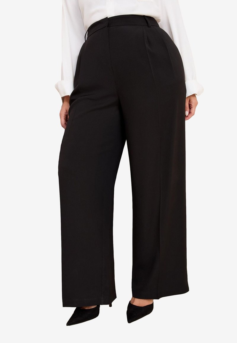 Donna che indossa pantaloni neri a vita alta e gamba larga abbinati a décolleté nere con punta a punta e una camicetta bianca a maniche lunghe.