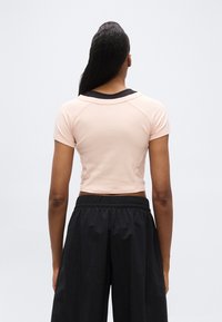 Γυναίκα με μακριά σκούρα μαλλιά που φοράει ανοιχτό ροζ κοντομάνικο κροπ τοπ πάνω από μαύρο πουκάμισο και φαρδιά μαύρη παντελόνια, εμφανιζόμενη από πίσω.