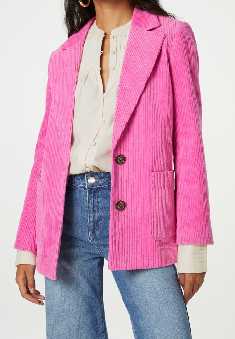 Giacca in corduroy rosa con rever a punta, due tasche frontali e due bottoni. Abbinata a una blusa crema e a jeans a gamba larga blu.