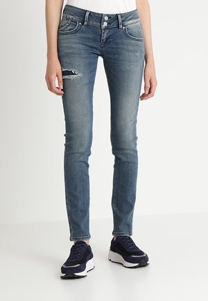 Kobieta w wyblakłych niebieskich obcisłych dżinsach z rozdarciem na lewej udzie i czarno-białych trampkach.