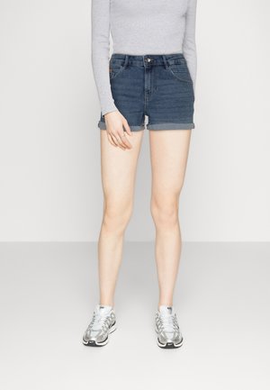 Shorts de mezclilla en azul medio con dobladillo enrollado, con bolsillos frontales y cierre de un solo botón, combinados con zapatillas deportivas plateadas.