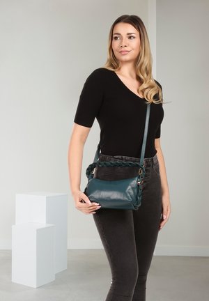 Jonge vrouw die een zwarte top en donkere jeans draagt, met een donkergroene leren handtas met gevlochten handvatriem, staande binnen.