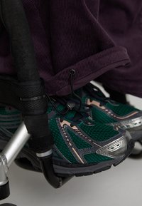 Pantofi sport verzi și gri cu țesătură din plasă și accente metalice, amplasați lângă un scaun cu rotile. Țesătura de catifea purpurie este parțial vizibilă deasupra.