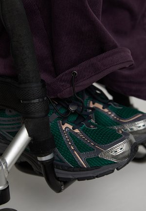 Zielone i szare buty sportowe z materiału siatkowego i metalicznymi akcentami, umieszczone obok wózka inwalidzkiego. Fioletowy materiał kordurowy częściowo widoczny powyżej.