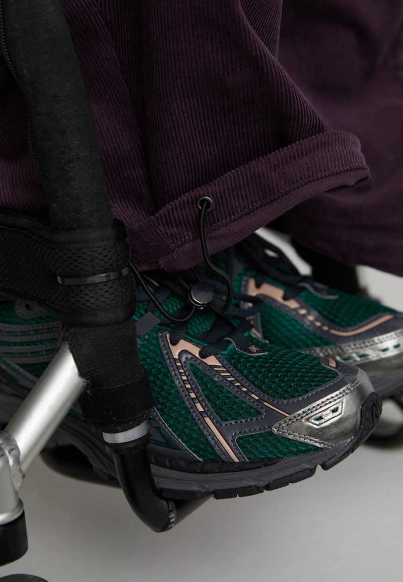 Pantofi sport verzi și gri cu țesătură din plasă și accente metalice, amplasați lângă un scaun cu rotile. Țesătura de catifea purpurie este parțial vizibilă deasupra.