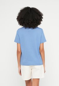 Kék színű pamut rövid ujjú póló, laza szabással, hátulnézetből. Világos színű rövidnadrággal párosítva.