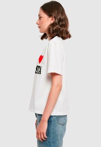 T-shirt en coton blanc avec un graphique de cœur rouge et un texte noir en gras. Manches courtes et coupe décontractée, associé à un jean bleu clair.