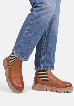 Brązowe skórzane botki sięgające kostki z paskami elastycznymi w paski po bokach oraz masywną jasnobrązową podeszwą, noszone z podwiniętymi niebieskimi dżinsami.