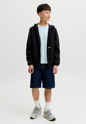 Junge steht und schaut nach rechts, trägt eine schwarze Kapuzenjacke, hellblaues Hemd, Jeansshorts, weiße Socken und graue Sneakers.
