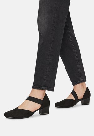 Černé semišové boty na širokém podpatku s v-řezem a elastickými pásky kolem kotníku, kombinované s tmavě modrými džínovými kalhotami.