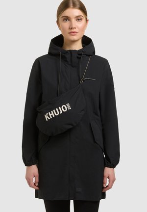 Žena v čiernom kapucňovom kabáte a čiernej crossbody taške s logom "KHUJO 2001" na prednej strane, stojaca pred jednoduchým svetlým pozadím.