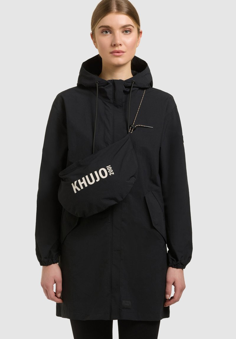 Mujer con un abrigo negro con capucha y un bolso cruzado negro con el logo "KHUJO 2001" en el frente, sobre un fondo liso y claro.