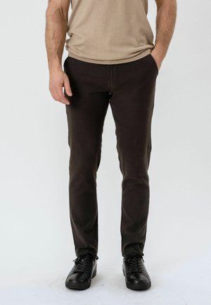 Mand iført slim-fit mørkebrune bukser, sorte læder sneakers og en beige skjorte med den ene hånd i lommen mod en hvid baggrund.