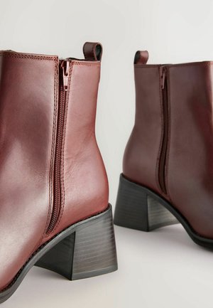 Brūni ādas potītes zābaki ar vidēja augstuma bloku papēdi, sānu iešuvēm un izšūtiem elementiem augšējā malā. Gluda tekstūra, elegants dizains.