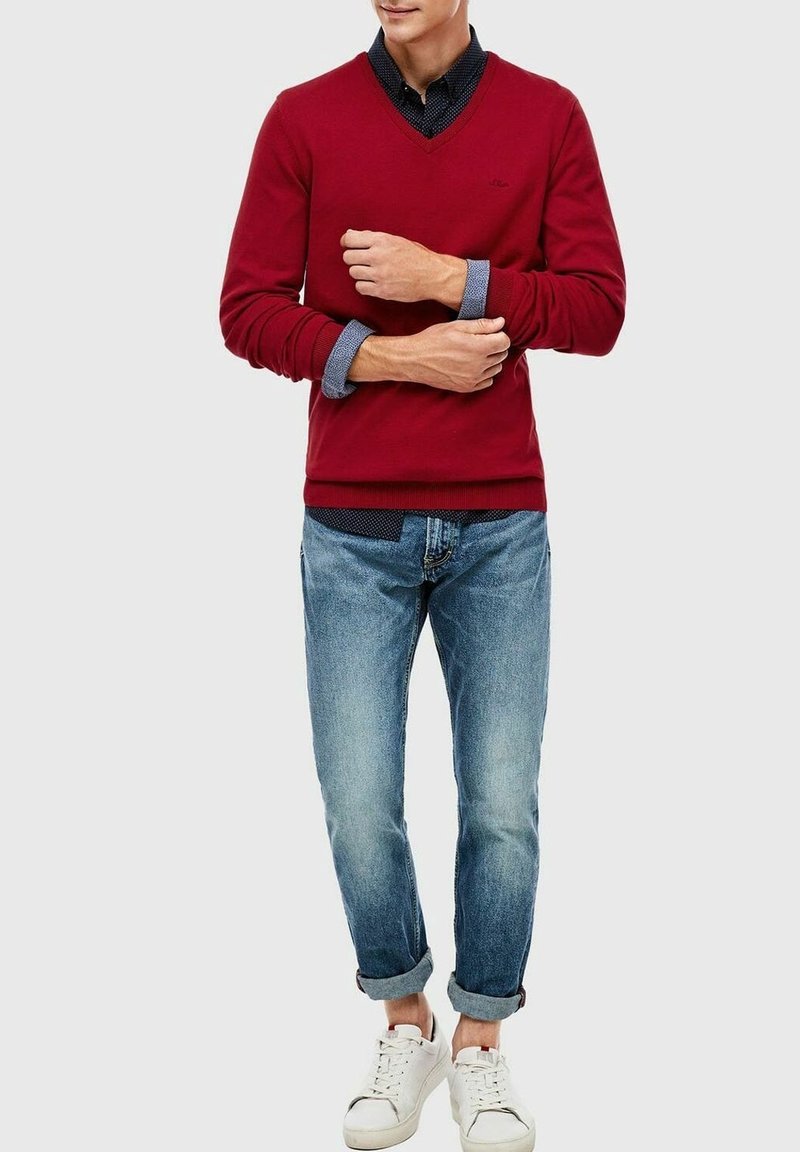 Pull rouge à col en V, superposé sur une chemise en chambray bleu. Associé à un jean bleu clair retroussé et des baskets blanches. Pas de motifs notables.