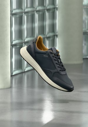 Schwarzer BOSS Sneaker mit weißer Sohle und beige Innenausstattung, der über einem polierten Betonboden nahe einer Glassteinwand schwebt.