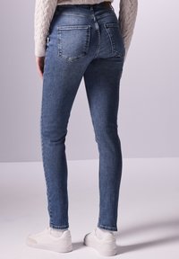 Jean en denim bleu skinny avec délavage, deux poches arrière et taille mi-haute. Porté avec des baskets blanches, montrant un look décontracté.