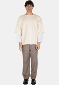T-shirt oversize beige sovrapposto a una camicia bianca a maniche lunghe, abbinato a pantaloni eleganti marroni e scarpe nere. Design semplice e contemporaneo.
