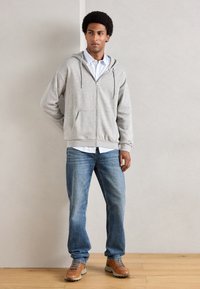 Jeune homme se tenant contre un mur clair, portant un sweat à capuche gris zippé, une chemise rayée, un jean bleu et des chaussures à lacets marron sur un sol en bois.