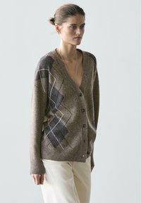 Kobieta ubrana w brązowy sweter na guziki z szaro-białymi wzorami w romby, w jasnobeżowych spodniach, stojąca na jednolitym tle.