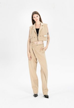 Joven mujer de pie con blazer corto sin mangas beige, pantalones de pierna ancha estilo cruzado, top negro y zapatos negros puntiagudos sobre fondo blanco.