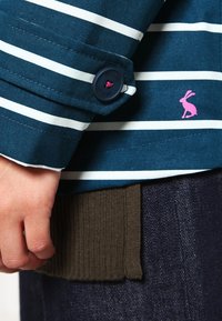 Käsi lepää tummansinisen ja valkoisen raidallisen pitkähihainen paidan napillisen kalvosimen vieressä, jossa on pieni vaaleanpunainen jänislogo, yllä tumman neulotun kankaan ja denimin päällä.