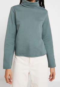 Person som bär en teal långärmad polotröja och off-white räfflade byxor, med flätat hår synligt vid axlarna.