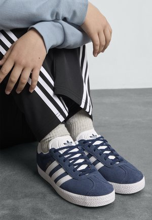 Baskets en daim bleu marine avec des accents rayés blancs, semelles en caoutchouc blanches et un design classique à lacets, portées avec des chaussettes grises et un pantalon noir.
