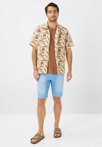 Camicetta a maniche corte beige con motivo floreale sopra una maglietta a collo tondo marrone, abbinata a pantaloni corti in denim azzurro chiaro e sandali scivolati marroni.