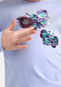 Lyse lilla langærmet skjorte med flerfarvede paliet sommerfugledesigner i blå, turkis og lilla. Blødt, tekstureret stof.