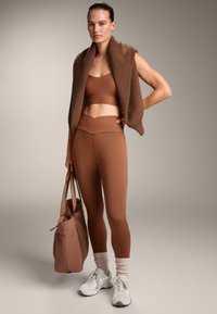 Barna sportos crop top és magas derekú leggings, barna kötött sál társaságában, valamint egy azonos színű puha táska, fehér sportcipők és világos zoknik.