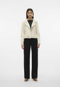 Veste en faux cuir couleur crème, crop, avec des zips argentés, associée à un pantalon large noir et des chaussures brunes. Texture lisse et coupe ajustée.