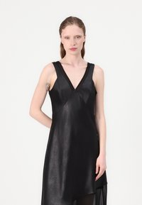 Black sleeveless dress with a V-neckline, featuring a textured fabric and asymmetrical hem. Has a cross-front design and sheer accents.