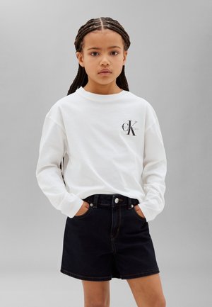 Młoda osoba z warkoczami, ubrana w białą koszulę Calvin Klein z długim rękawem i czarne jeansowe szorty, stojąca z rękami w kieszeniach.