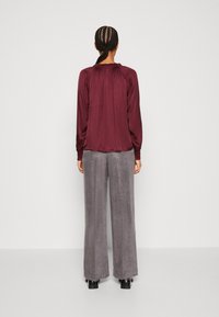 Burgunderfarbene Bluse mit langen Ärmeln und gerafftem Ausschnitt, kombiniert mit grauen, weiten Hosen aus einem weichen, strukturierten Stoff. Schwarze Absatzschuhe.