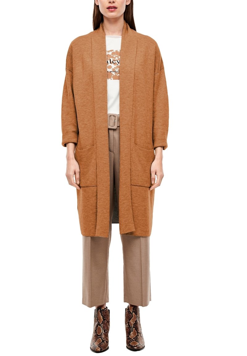 Světle hnědý kardigan s otevřeným předním dílem, spadlými rameny, dvěma předními kapsami a žebrovanou texturou, nošený přes bílé tričko a béžové kalhoty.