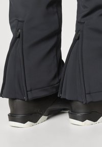 Μαύρο αδιάβροχο παντελόνι με φερμουάρ στους αστραγάλους, σε συνδυασμό με μαύρες μπότες με υφή στη σόλα και λευκές λεπτομέρειες.