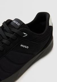 Chaussure de sport noire avec une tige texturée comprenant des panneaux en mesh, des détails de laçage et une semelle en caoutchouc. Logo "HUGO" sur la languette et le côté.