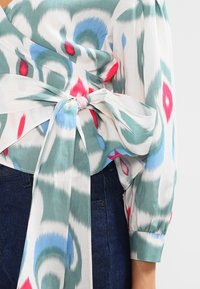 Välja, mönstrad blus i lätt tyg med gröna, blå, röda och vita färger. Med knytning i midjan och långa flödande ärmar.