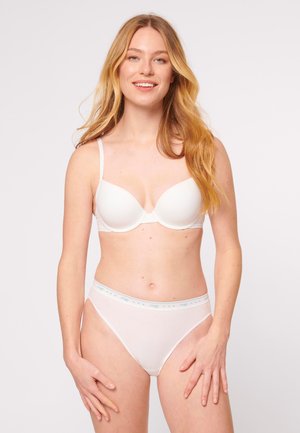 Donna con capelli biondi lunghi che sorride, indossando un reggiseno bianco e mutandine a vita alta, in piedi contro uno sfondo chiaro e uniforme.