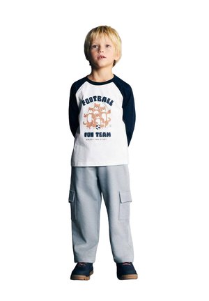 Blond chlapec stojí, má na sebe bielo-námornícku tričko s dlhými rukávmi s kreslenou témou futbalu a sivé cargo nohavice s čiernymi topánkami.