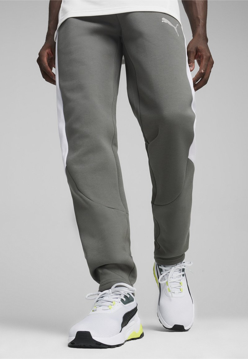 Szare dresy Puma z białymi panelami bocznymi, charakteryzujące się gładką teksturą i zwężanym krojem. W połączeniu z białymi i zielonymi butami do biegania.