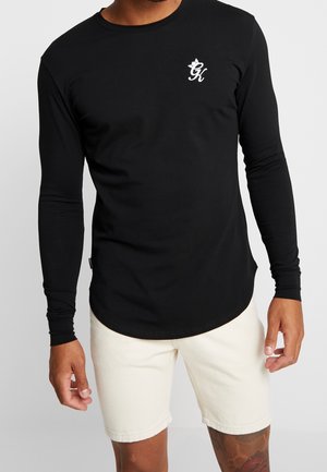 Schwarzes Langarmshirt aus weichem Stoff, mit einem weißen Logo auf der Brust, abgerundetem Saum und schmalen Ärmeln. Kombiniert mit hellen Shorts.