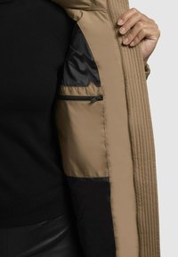 Beige gewatteerde jas binnenkant met een gladde zwarte voering en een zijzak met rits. Textuursteek langs de rand voegt detail toe.
