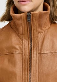 Donna che indossa una giacca di pelle color beige con un alto colletto a coste e una cerniera centrale, mostrata dalla mento fino a metà petto.