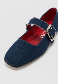 Chaussure Mary Jane en denim bleu avec un bout carré, une sangle effilochée et une boucle en argent, avec une doublure intérieure rouge et une semelle plate noire.