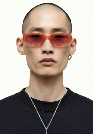 Personne aux cheveux courts portant des lunettes de soleil teintées orange, un anneau argenté à l'oreille, une chaîne argentée autour du cou et un t-shirt noir à col ras du cou.