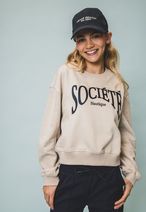 Beige sweatshirt met "SOCIÉTÉ Nautique" in marineblauwe letters, gecombineerd met marineblauwe joggingbroek met trekkoord en een zwarte pet met de tekst "MIAMI BEACH".