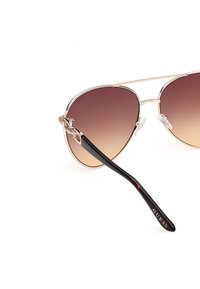 Gafas de sol estilo aviador con lentes marrones degradados, montura de metal dorado y patillas de carey negro. Nombre de la marca 'GUESS' en la patilla.